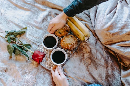 Plateau avec deux tasses de café, des muffins, des bananes et une rose sur un lit en velours. Ambiance romantique.